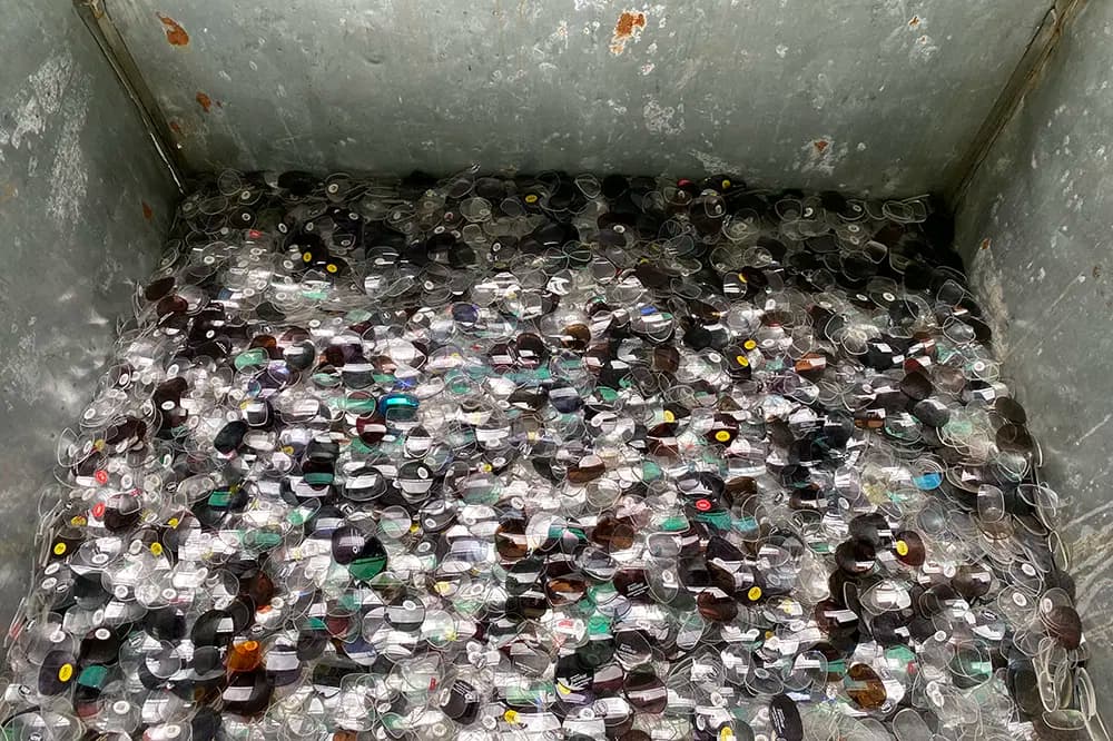 Thousands of discarded eyeglass and sunglass lenses filling a large, weathered metal industrial bin.