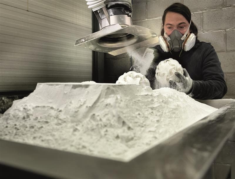Technician in a respirator cleans an intricate 3D-printed sphere from a bed of powder.